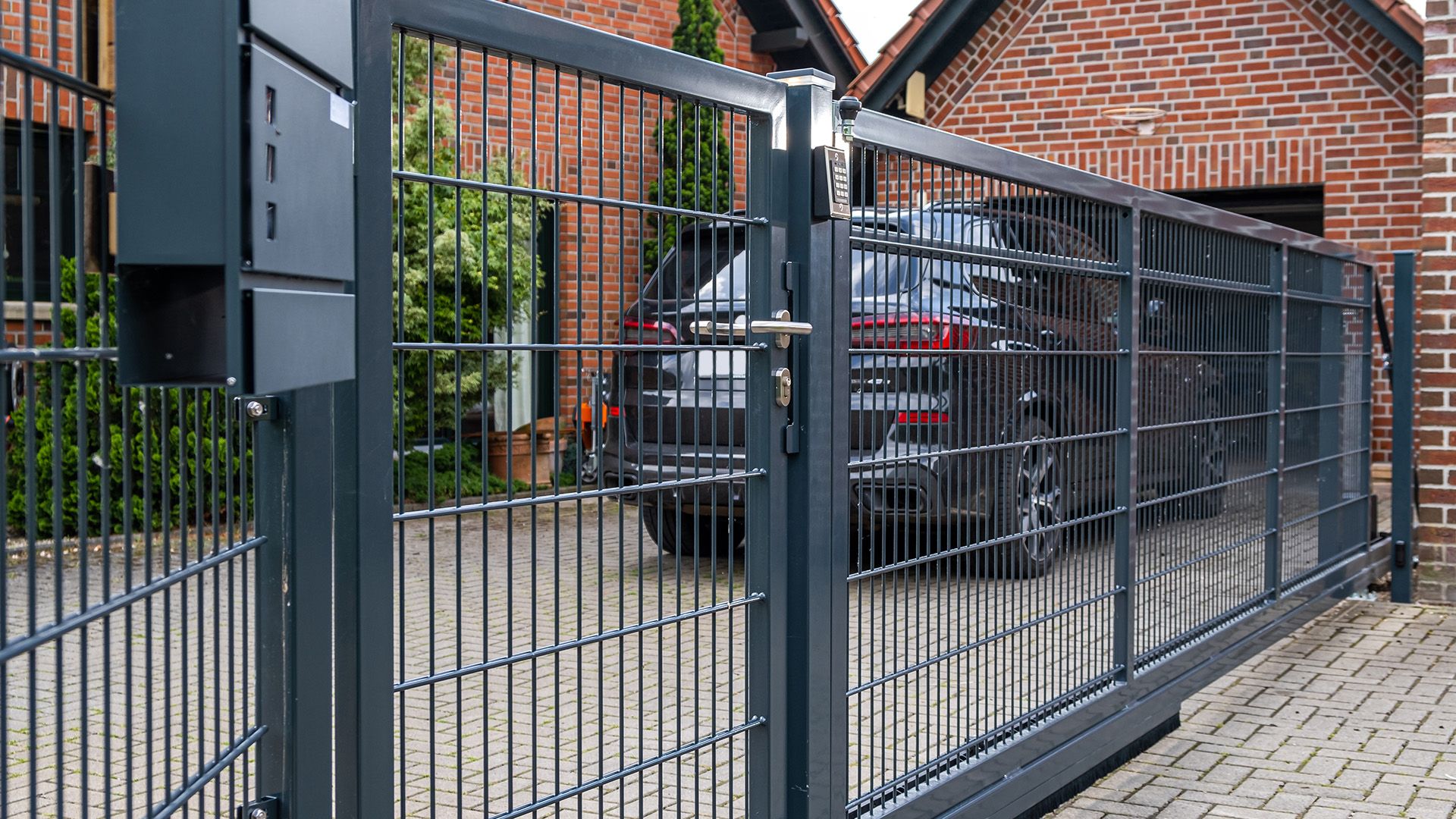 Eine Schiebetoranlage an einer Einfahrt mit einem Klinker-Wohnhaus und einem Auto im Hintergrund.