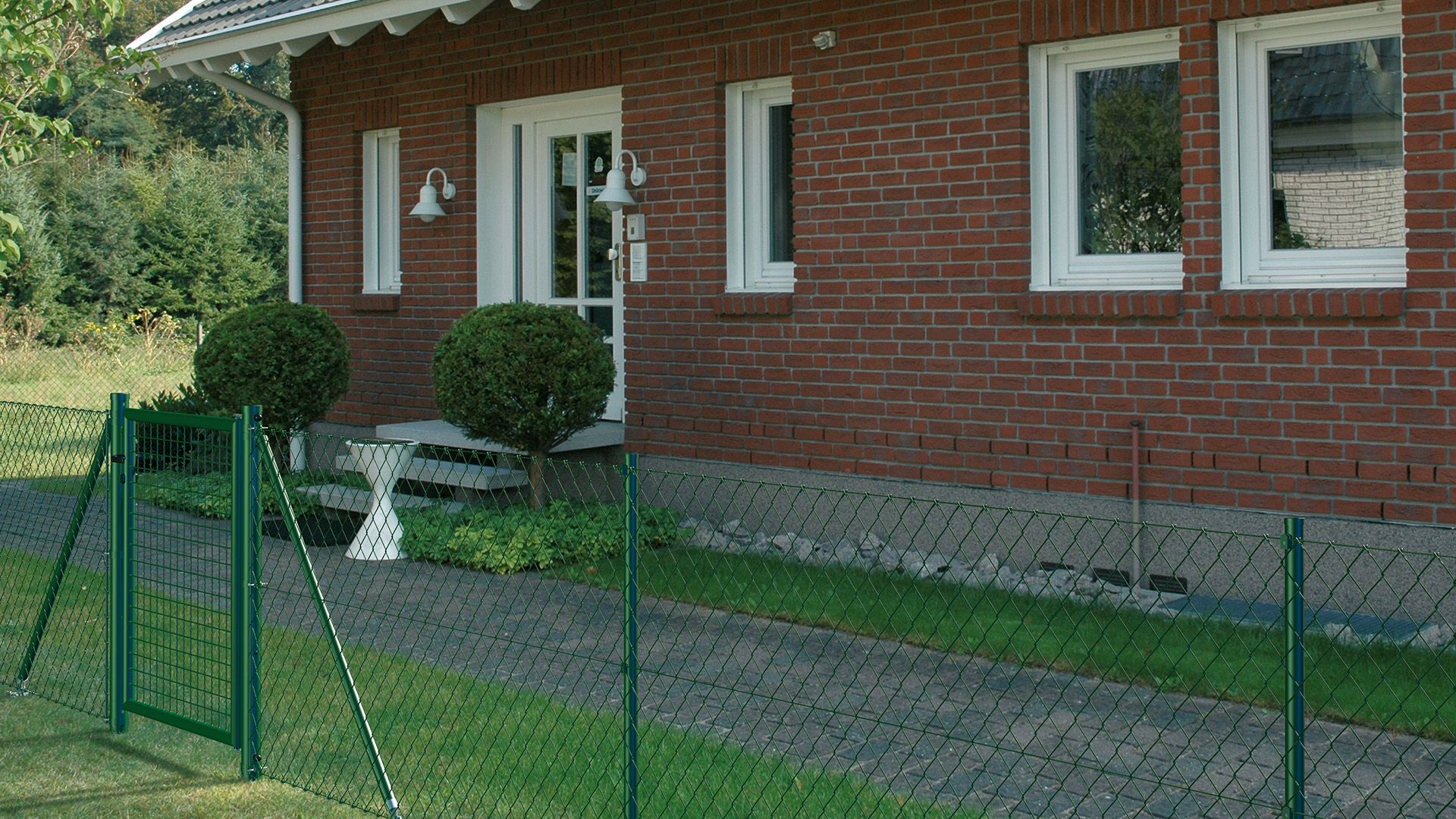 Maschendrahtzaun mit Wellengitter-Einzeltor in RAL 6005 Grün mit einem Haus und Gartenweg im Hintergrund.